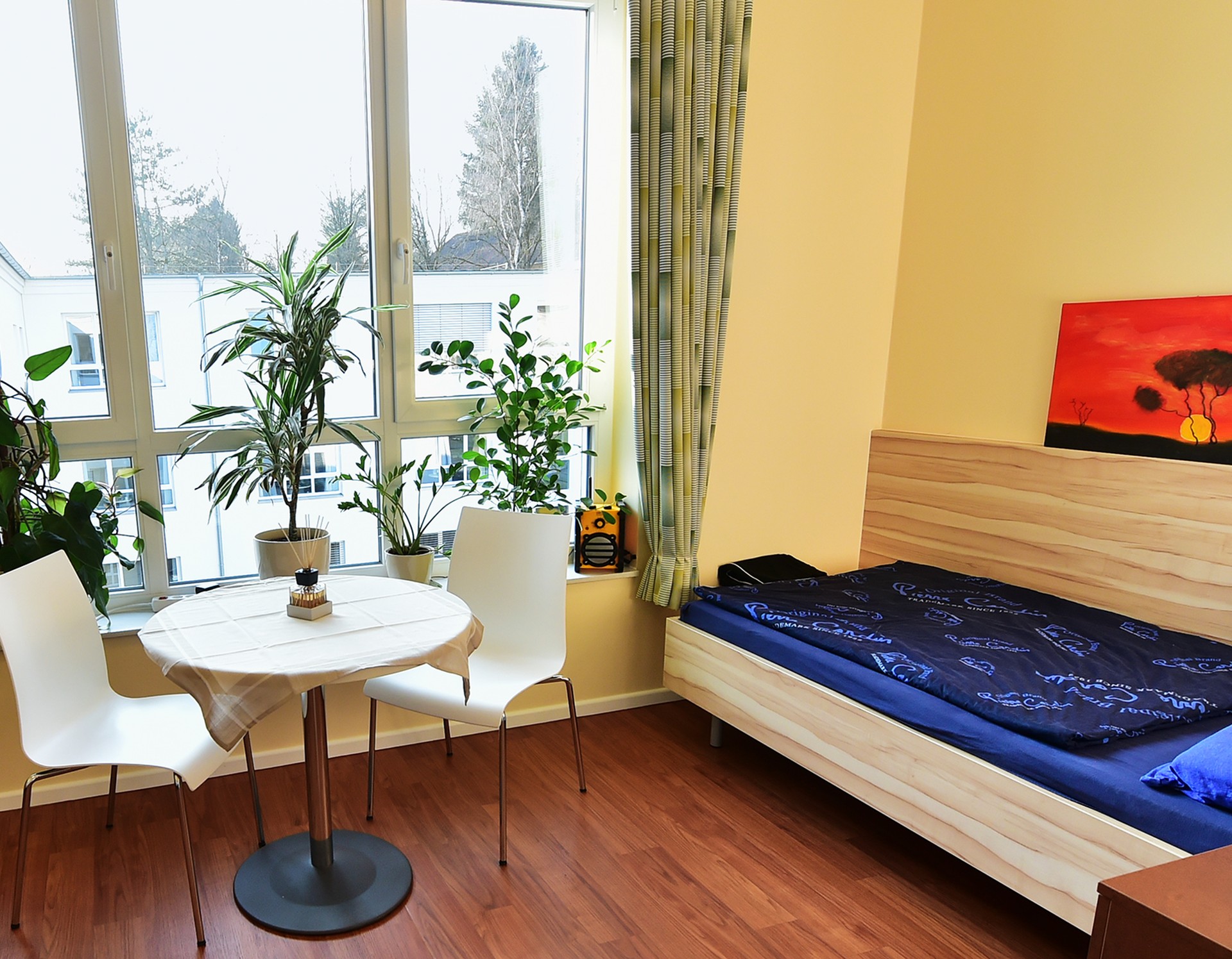 Ein Einzelzimmer der Sucht-Reha mit einem Bett, einer Sitzecke, einem großen Fenster und Bepflanzung